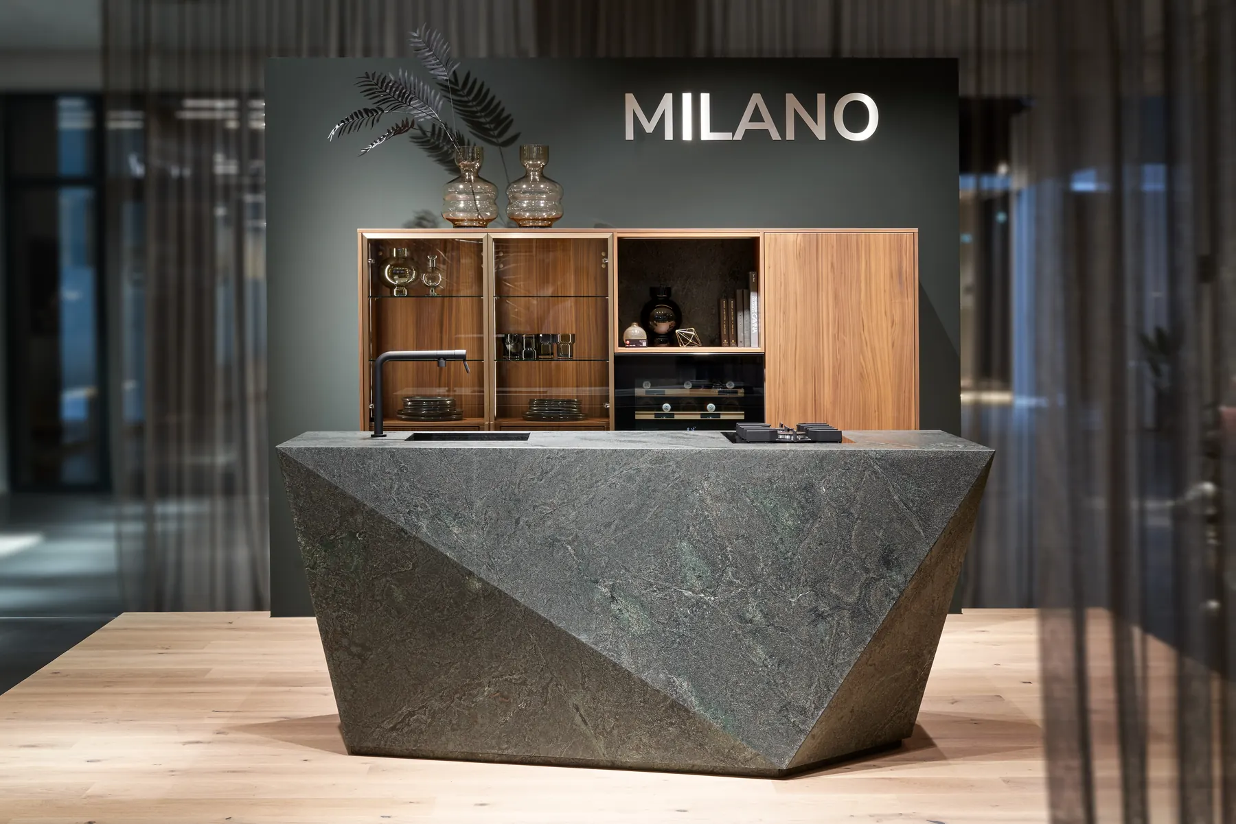 Kochinsel Milano mit facettiertem Korpus aus grünem Naturstein vor Vitrinen in Nussbaum