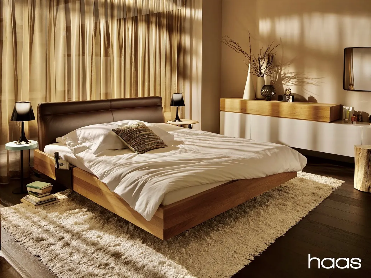 Bett mit braunem Lederkopfteil und Bettrahmen aus Eiche vor lichtdurchflutetem Vorhang, daneben ein Sideboard