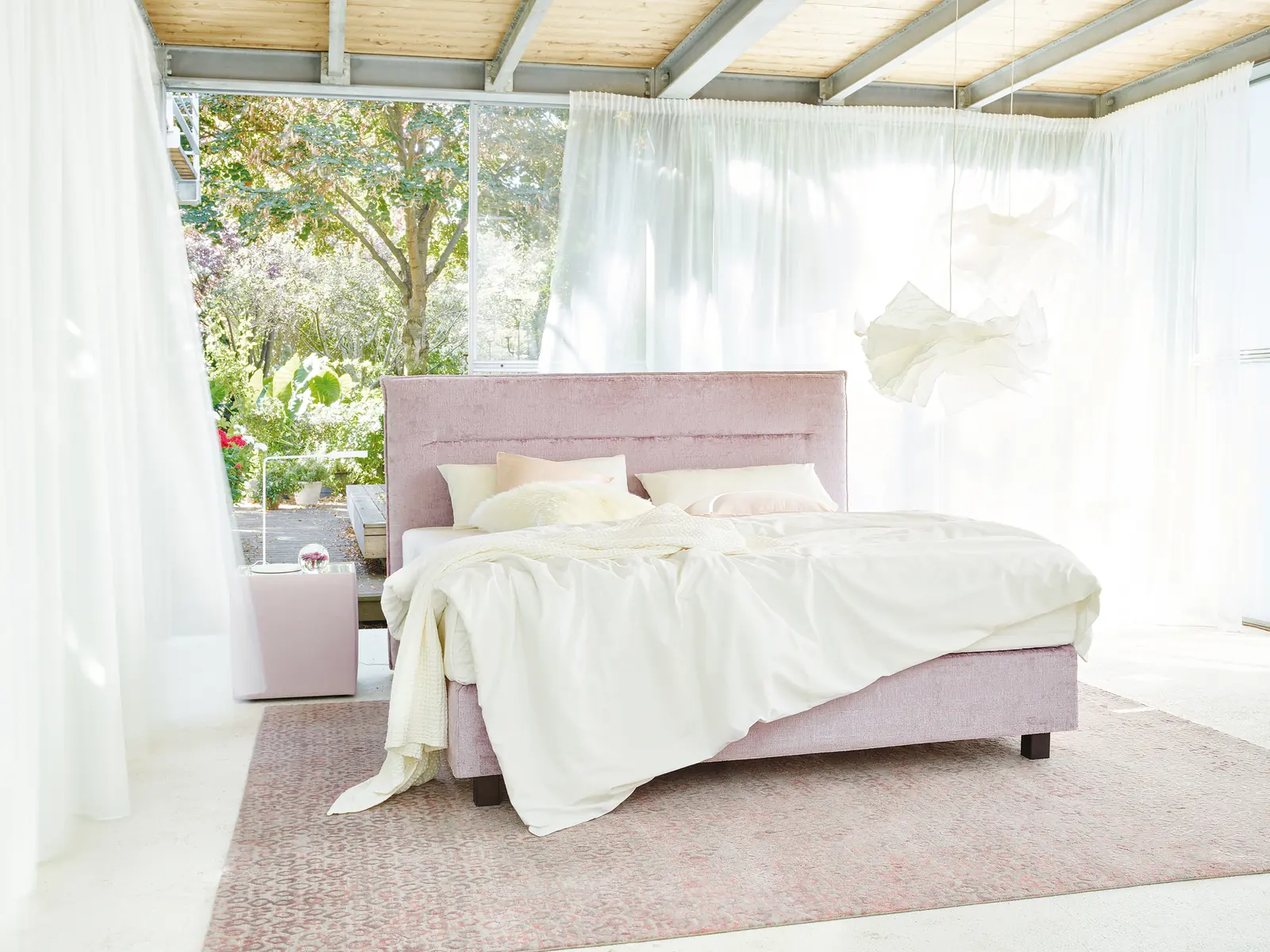 Boxspringbett Capri in Rosé mit hellem Bettzeug vor bodentiefen Fenstern