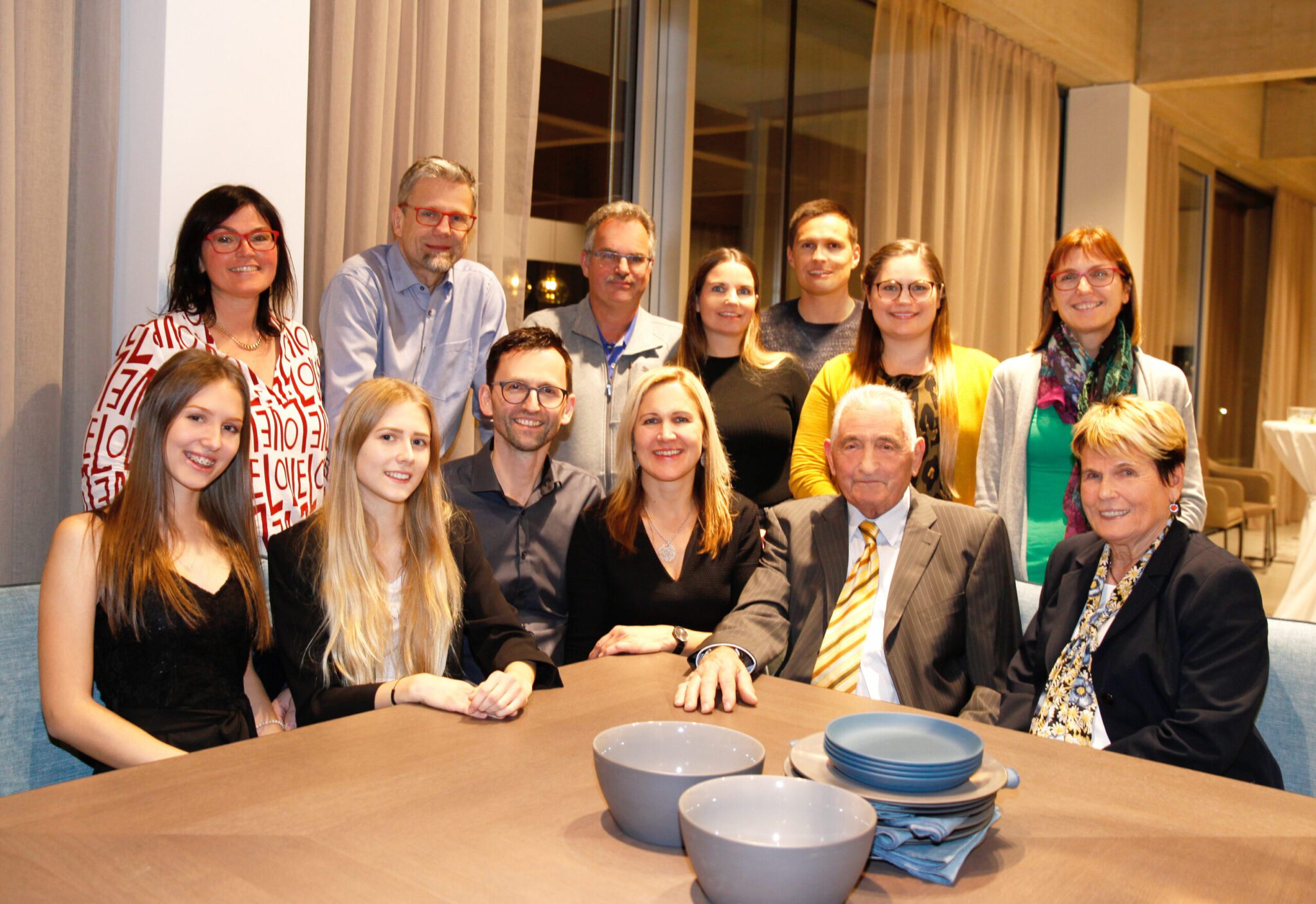 Gruppenfoto zur Eröffnung 2019: Gäste sitzen und stehen gemeinsam an einem Esstisch im Schauraum
