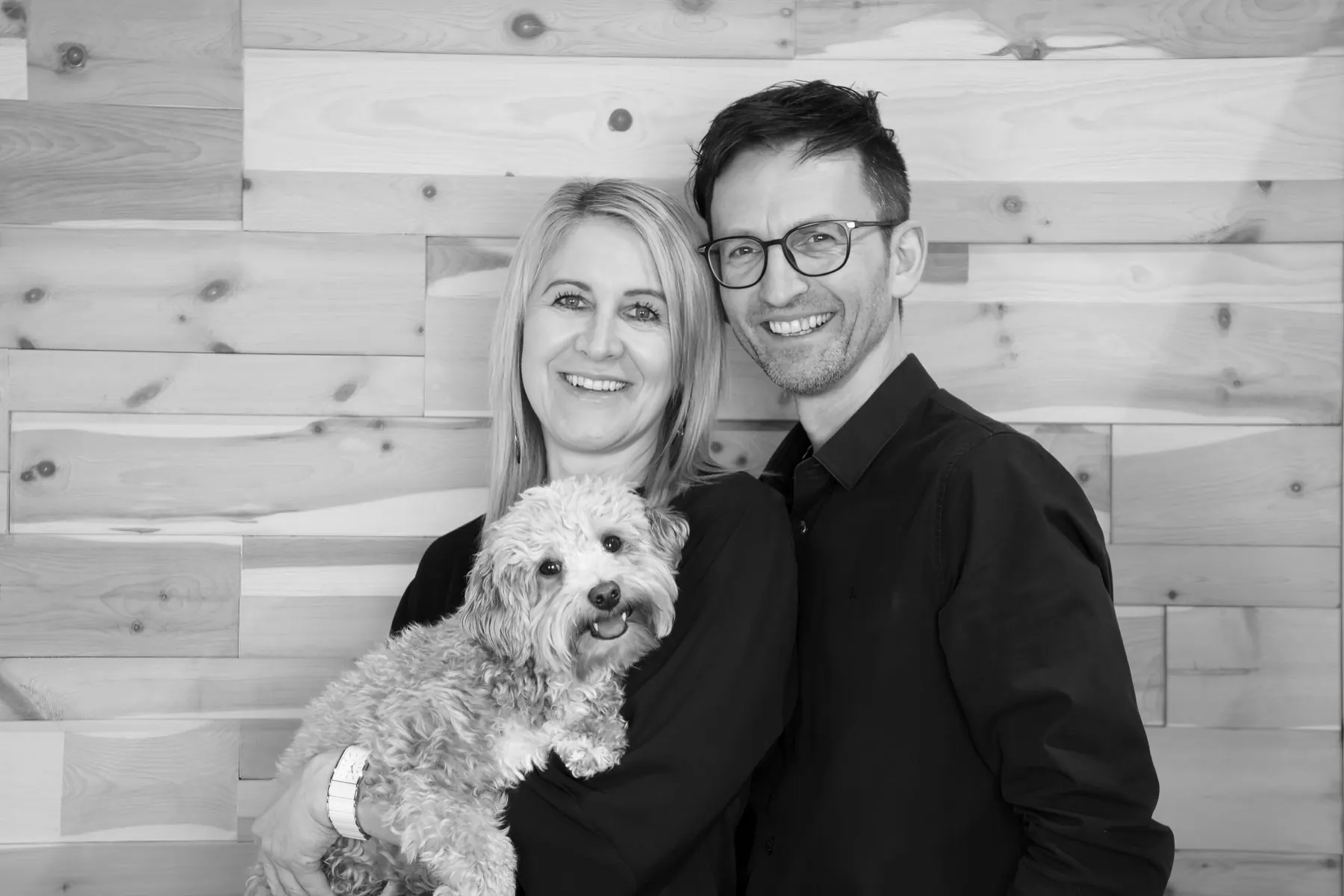 Schwarz-Weiß-Porträt des Inhaberpaars mit Hund vor einer Massivholzwand